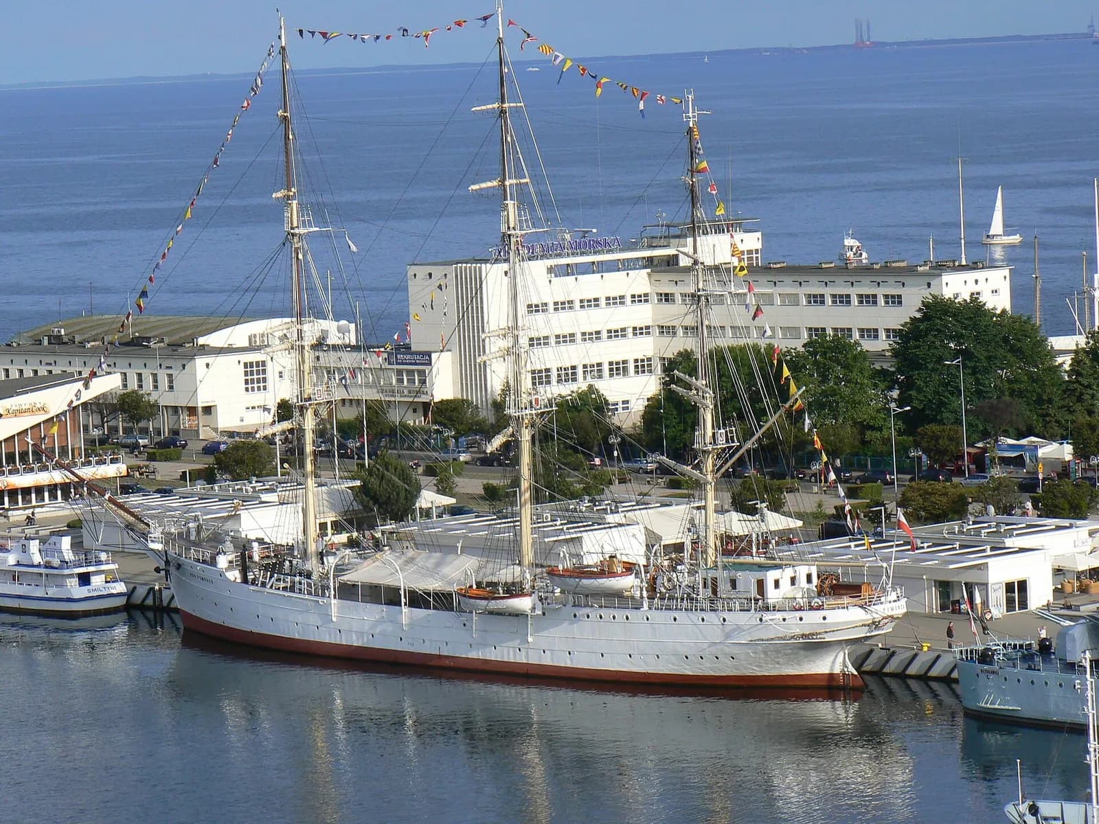 A large, white, multi-masted sailing ship with red trim docked in a harbor, adorned with colorful flags, with white buildings and serene blue water in the background.