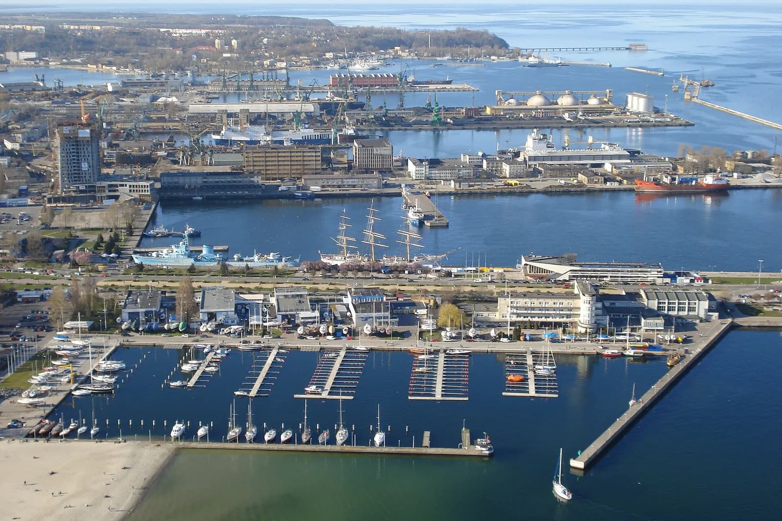 A modern cityscape featuring tall, sleek buildings with varying architectural styles, some reflecting the blue sky and clouds, situated along a body of water with small boats docked nearby.
